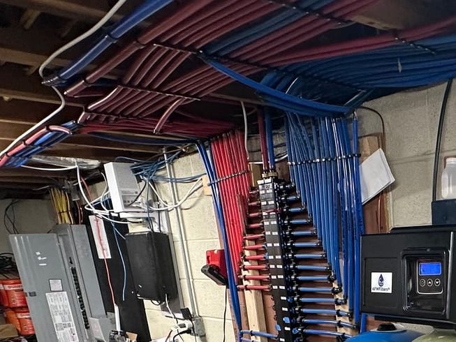 Basement manifold with blue and red PEX supply lines running up to the ceiling joists and a white drain stack