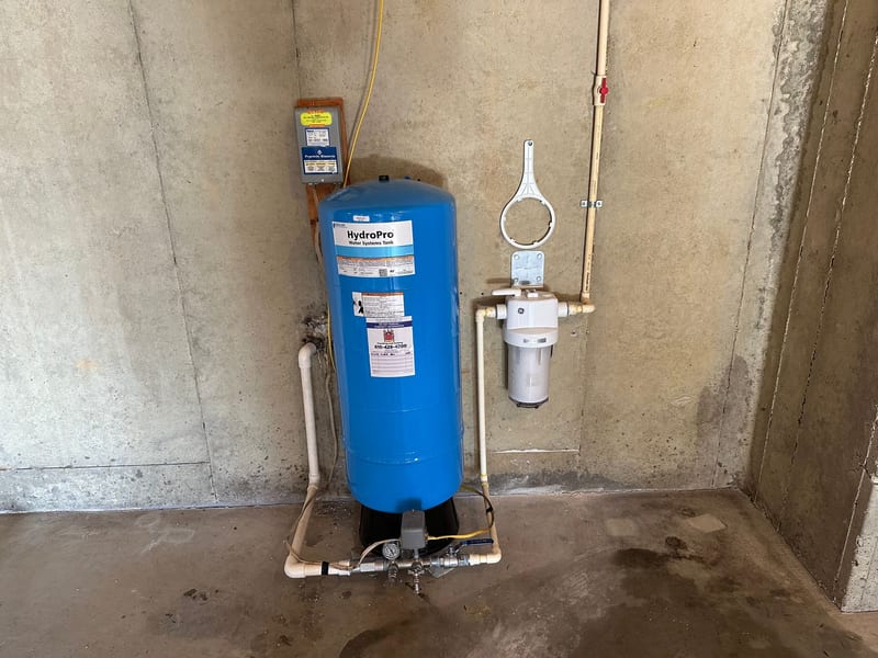 Blue well pressure tank with a pressure gauge, valves and white piping on a basement floor