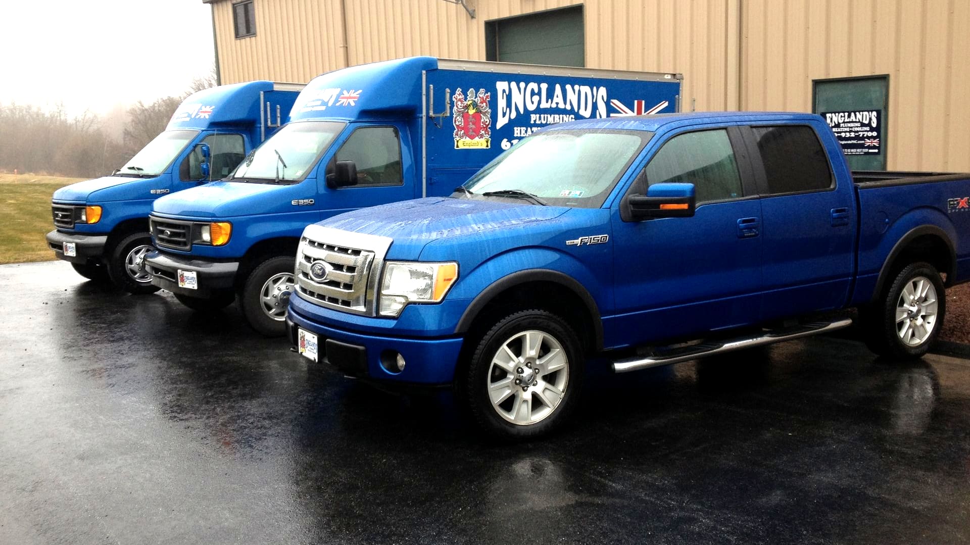 England's Plumbing and Heating fleet: two blue box trucks lettered ENGLAND'S Plumbing Heating Cooling with the family crest, and a blue pickup, parked in front of the shop