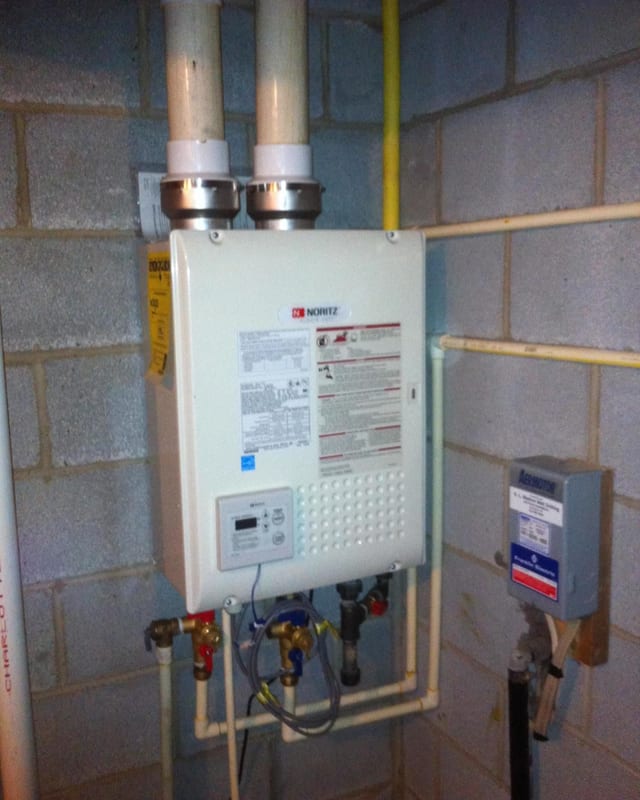 Tankless water heater on a cinder block wall with twin white vent pipes and a gas line